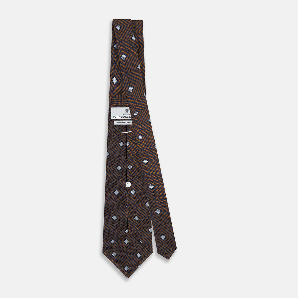 The Turnbull & Asser UK Tomorrow Never Dies Seven-Fold Silk Tie, as seen on James Bond, features a navy blue and brown geometric diamond pattern with silver squares at the center, set on a light background.