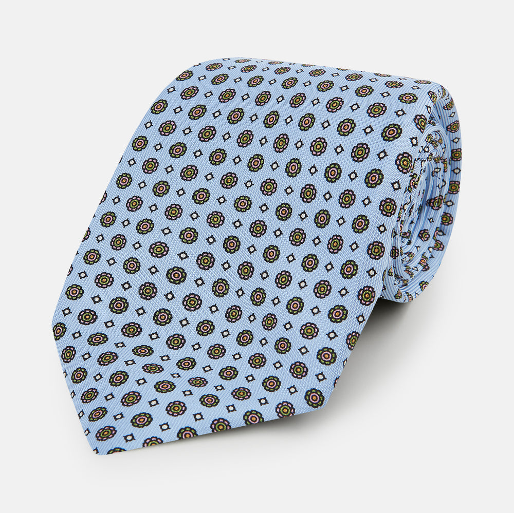 The Turnbull & Asser UK Great Gatsby Seven-Fold Silk Sky Tie, as seen on Jay Gatsby, is a light blue silk tie with a repeating floral medallion pattern in black, yellow, and red circles, shown rolled on a white background.