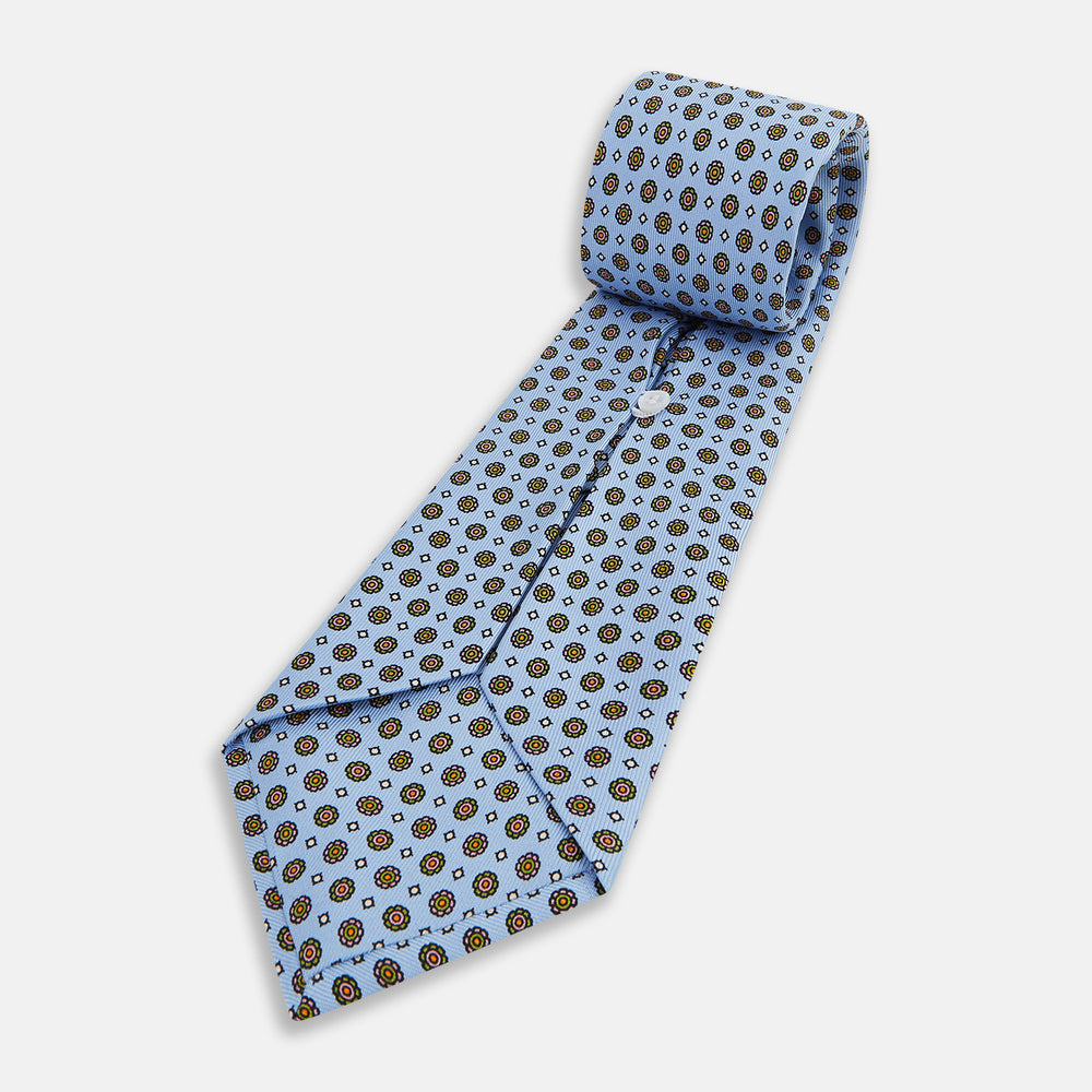 The Turnbull & Asser UK Great Gatsby Seven-Fold Silk Sky Tie, as seen on Jay Gatsby, is a light blue silk tie with a repeating floral medallion pattern in black, yellow, and red circles, shown rolled on a white background.