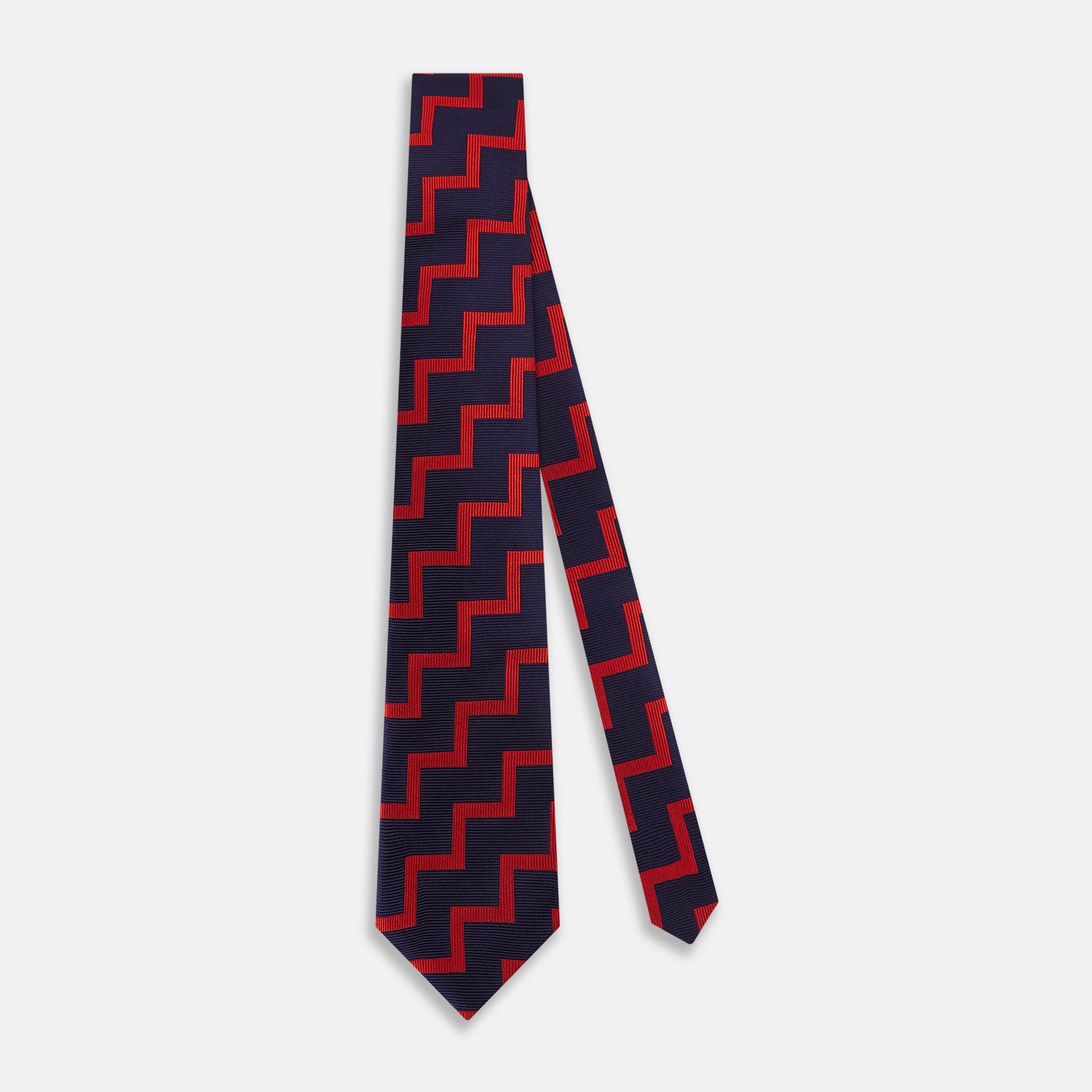 View 2 of product The Turnbull & Asser UK Seven-Fold Silk Red and Navy Geometric Tie is displayed rolled with its tip forward, showcasing its bold navy and red geometric pattern against a plain white background.