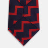 View 3 of product The Turnbull & Asser UK Seven-Fold Silk Red and Navy Geometric Tie is displayed rolled with its tip forward, showcasing its bold navy and red geometric pattern against a plain white background.