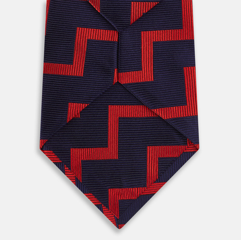 The Turnbull & Asser UK Seven-Fold Silk Red and Navy Geometric Tie is displayed rolled with its tip forward, showcasing its bold navy and red geometric pattern against a plain white background.