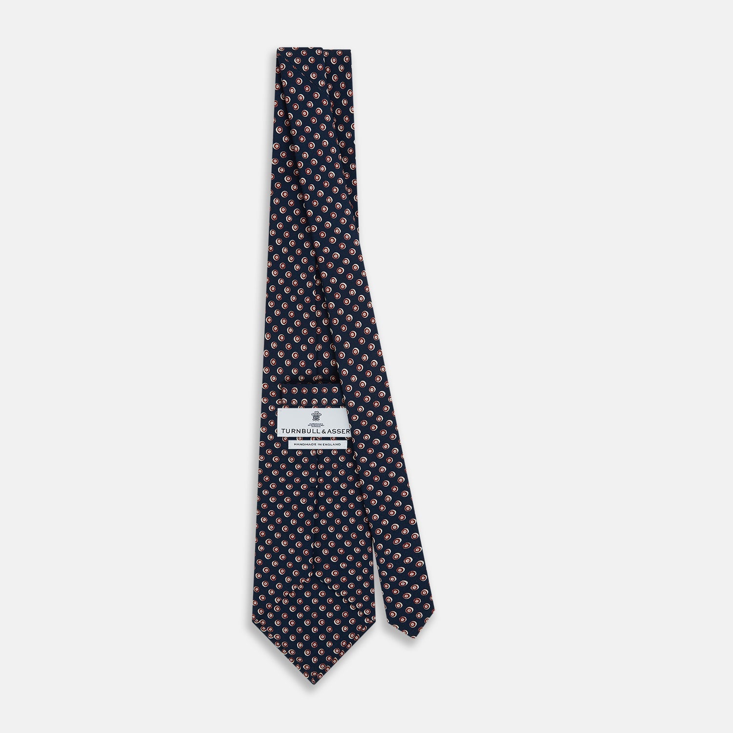 View 4 of product The Turnbull & Asser Navy and Red Circle Foulard Silk Tie, featuring a repeating red and white circular motif, is neatly rolled to display classic British craftsmanship.