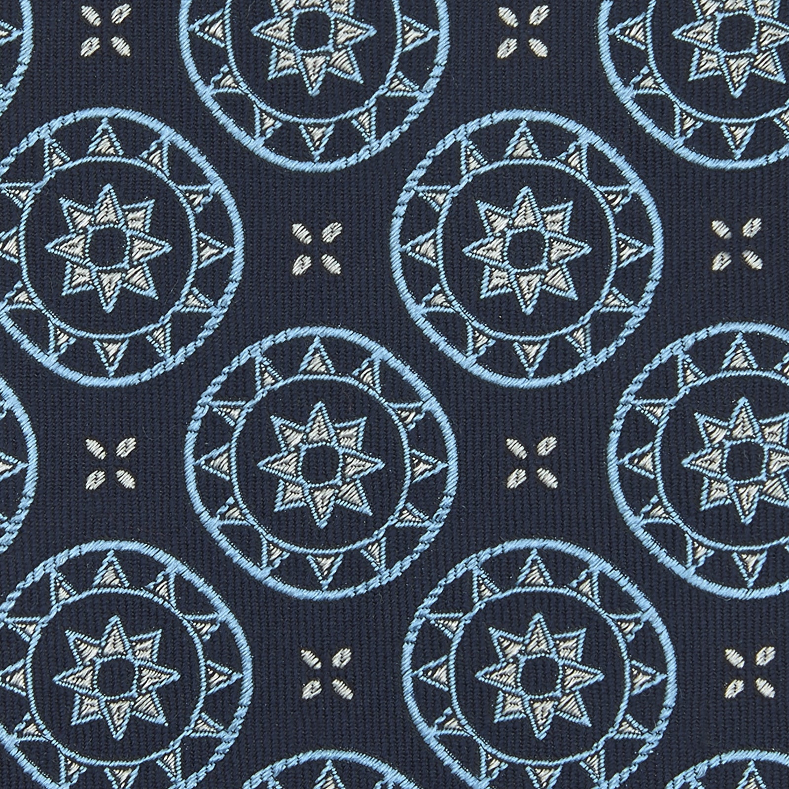 View 4 of product The Jacquard Silk Geometric Tie by Turnbull & Asser features a navy blue base with light blue and white star and circle motifs, shown rolled on a white background.