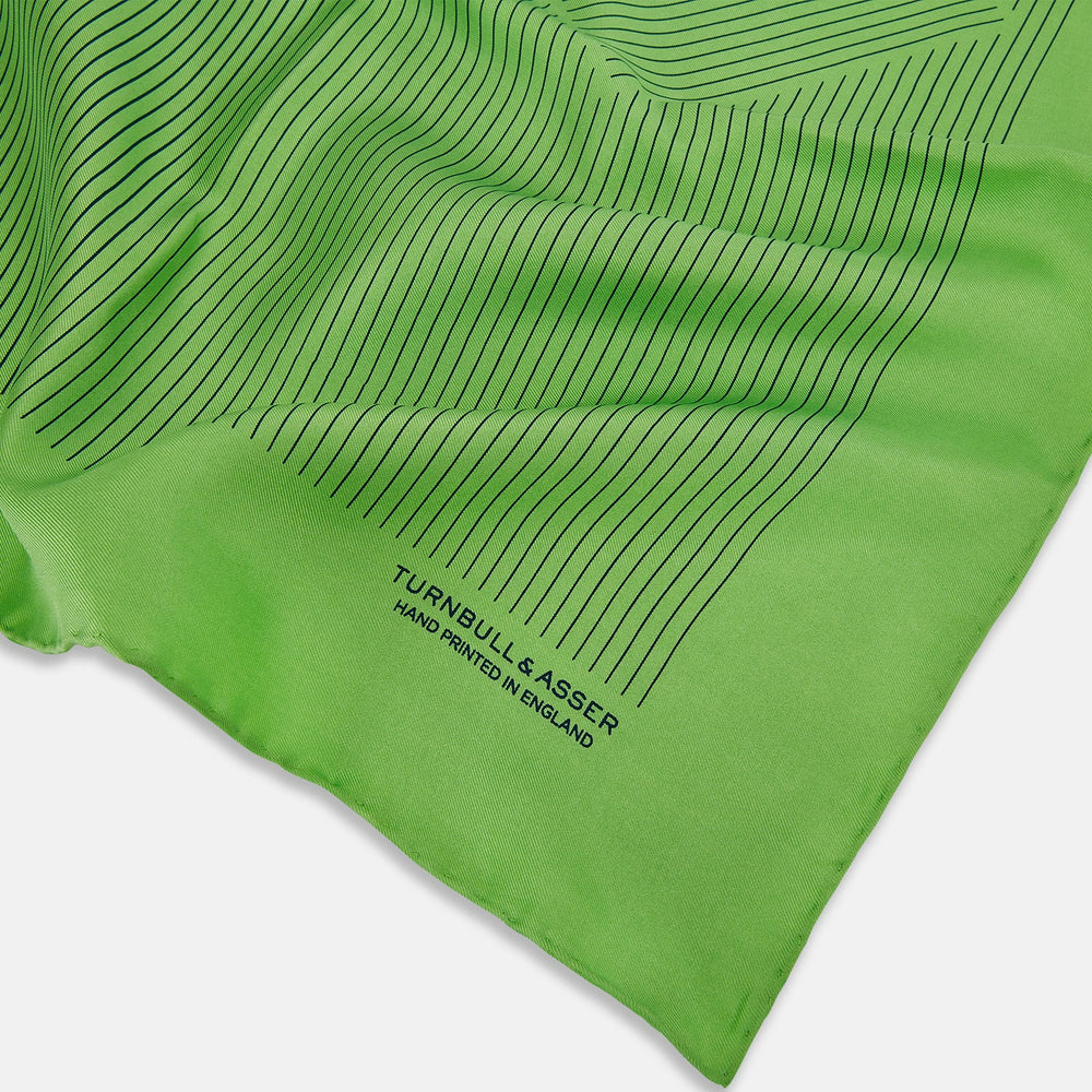The Fern Green Stripe Silk Pocket Square by Turnbull & Asser features thin black vertical stripes, celebrates British heritage, and is inscribed “TURNBULL & ASSER HAND PRINTED IN ENGLAND” in one corner; one corner is folded inward.