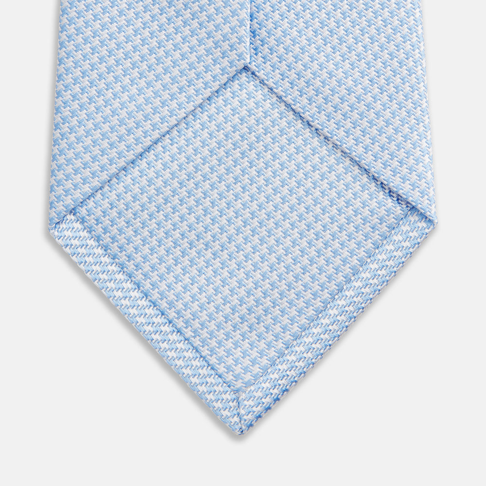 The HOUNDSTOOTH WHITE RIVER TIE by Turnbull & Asser features a light blue and white textured pattern, making it an essential accessory for elegant looks, shown beautifully rolled on a plain white background.