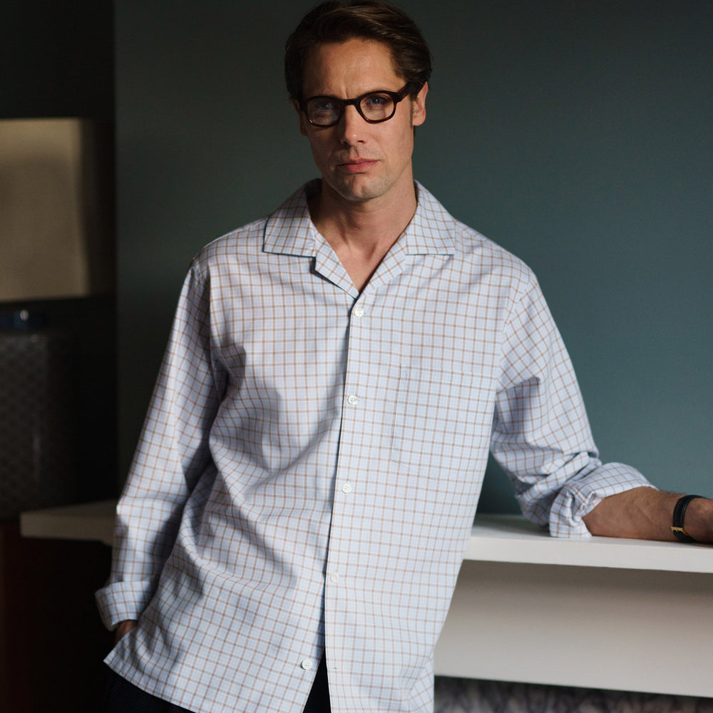 A man wearing glasses and a Pale Blue Multi Check Cashmerello Holiday Fit Winnington Shirt by Turnbull & Asser leans casually against a white countertop, looking at the camera with a neutral expression.
