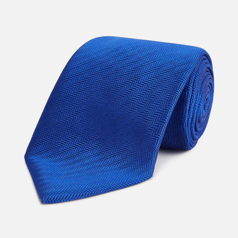 The Turnbull & Asser Plain Herringbone Cruise Tie in bright blue, shown rolled up on a white background, features a subtle chevron pattern—an essential finishing touch for your smart look.