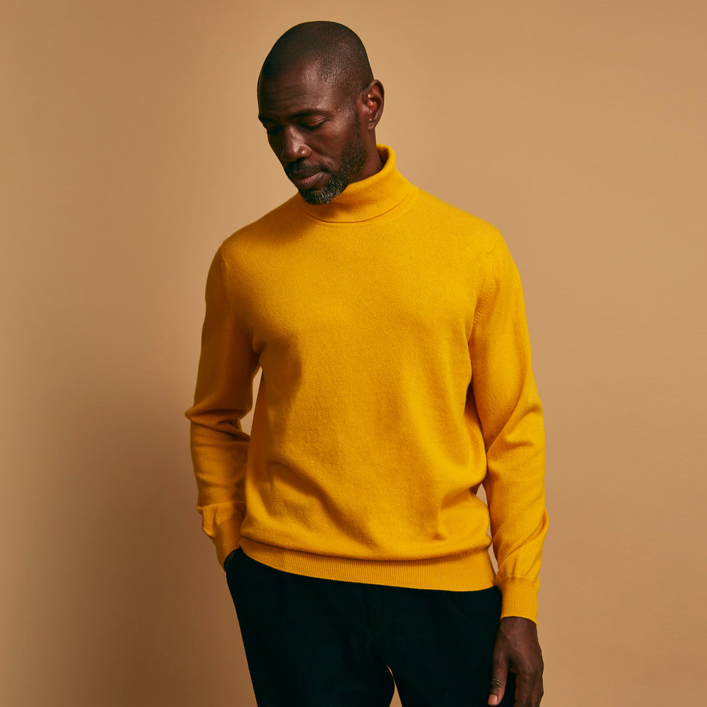 A man with short hair and a beard wears the Gold Aubrey Cashmere Roll Neck Jumper by Turnbull & Asser with dark pants, standing against a plain beige background and calmly looking at the camera, highlighting this stylish autumn sweater look.