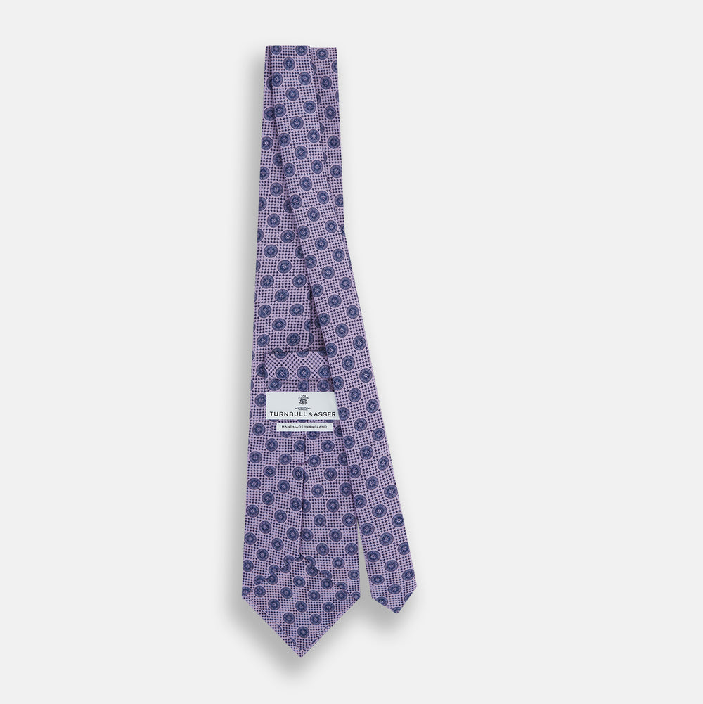 The Turnbull & Asser Lilac Medallion Printed Silk Tie is crafted from English silk with a geometric motif of small blue circles and a subtle checkered background.
