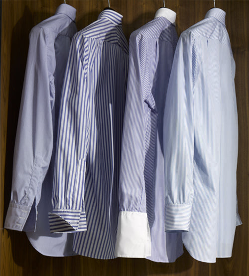 Five long-sleeved dress shirts in various patterns and shades of blue hang neatly side by side on a wooden background, with the stripes and cuffs prominently displayed.