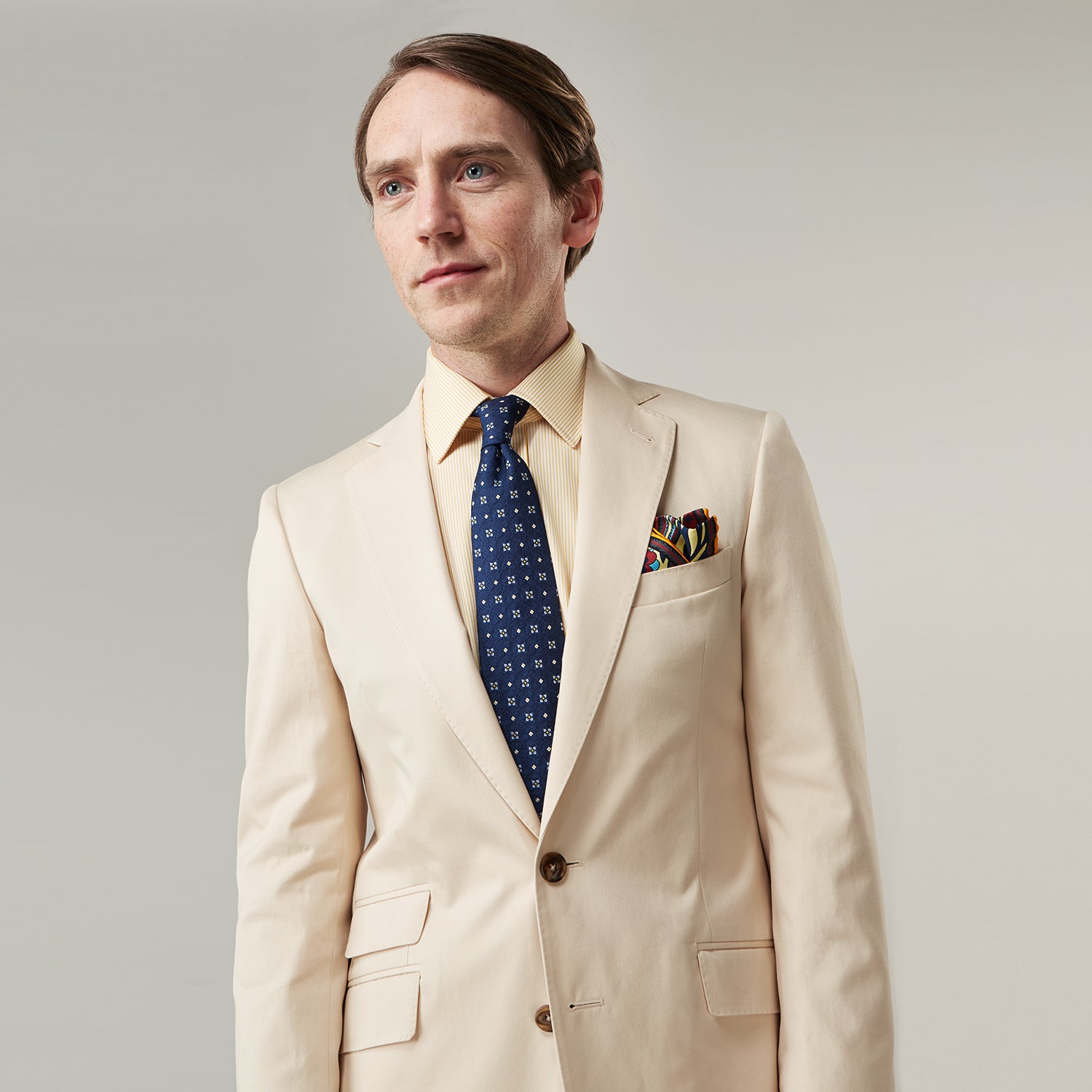 View 7 of product A man in a Turnbull & Asser Beige Cotton Barrington Blazer, light yellow shirt, blue patterned tie, and dark trousers stands against a plain light background, with a pocket square visible and a neutral expression.
