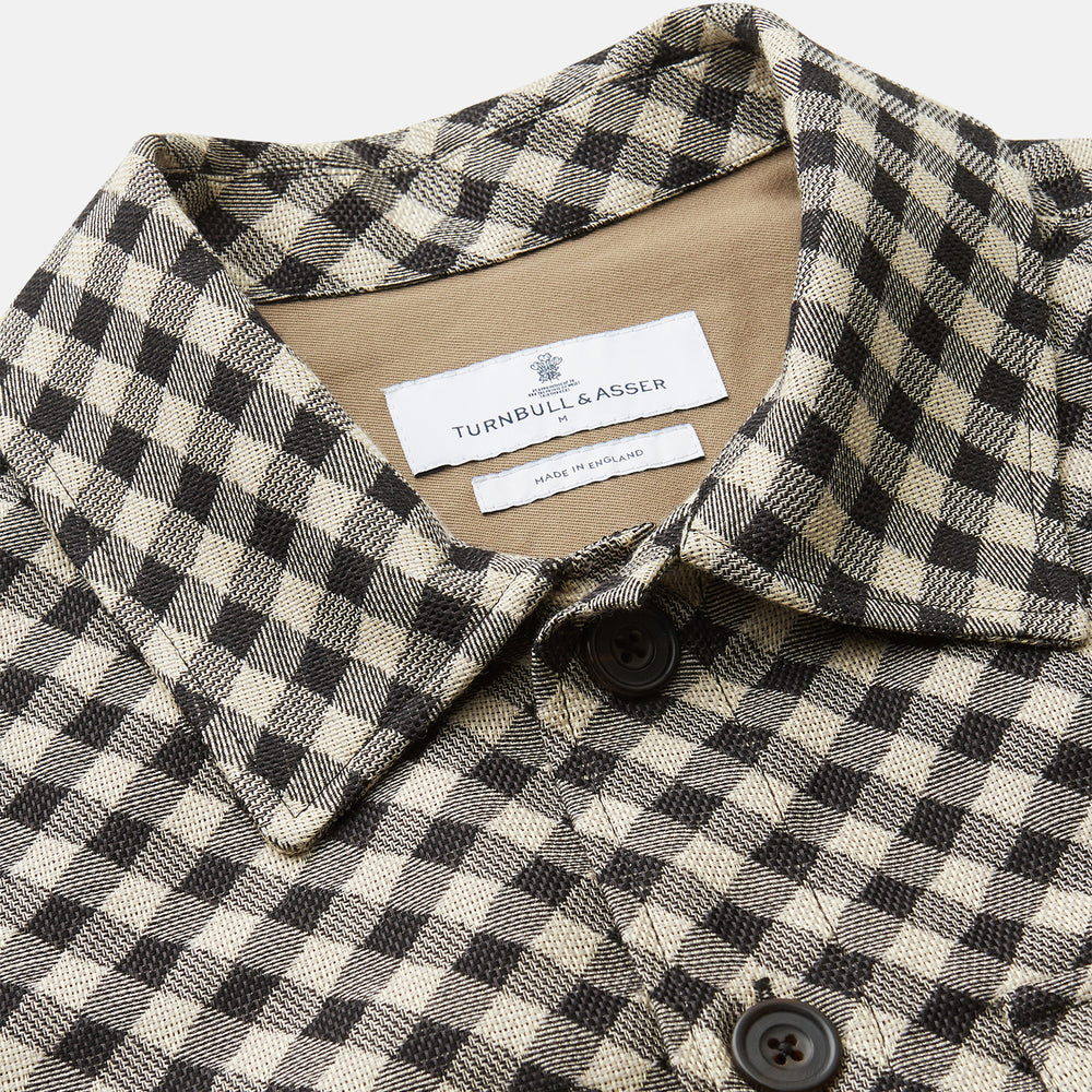 The Turnbull & Asser Black Gingham Check Wool Blend Somerset Overshirt features long sleeves, two chest flap pockets with buttons, and a black-and-beige check pattern. Shown laid flat on a white background; ideal as transitional outerwear.