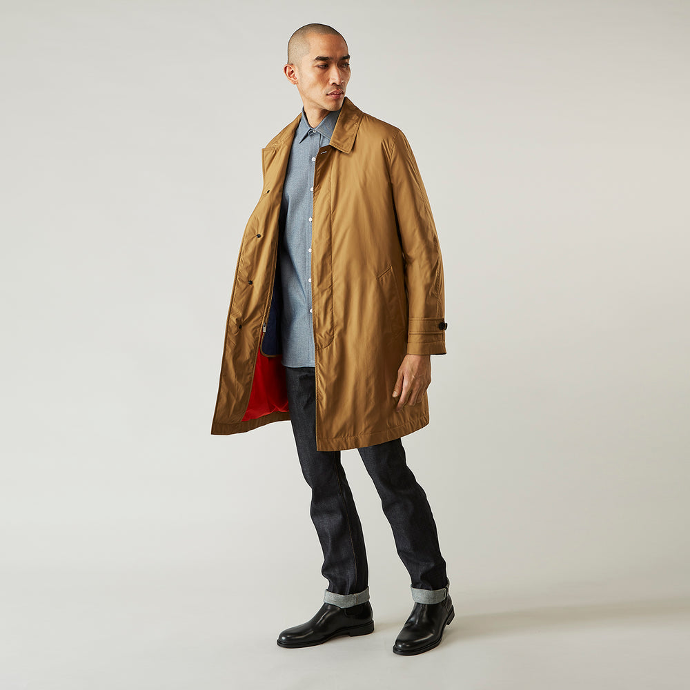 A person wears the Turnbull & Asser Camel Quintern Sealup Raincoat with Detachable Liner, paired with dark pants, standing against a plain light gray background, showing the sleek technical rainwear style with one hand in a pocket.