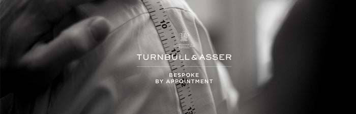 A close-up of a tailor measuring a persons shoulder with a measuring tape, overlaid with the text Turnbull & Asser Bespoke By Appointment in white letters.