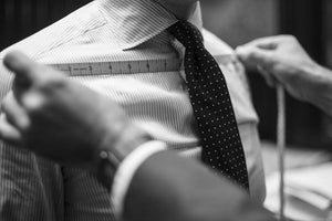 A tailor measures a mans shoulder width with a ruler while the man wears a striped dress shirt and polka dot tie. The image is in black and white.