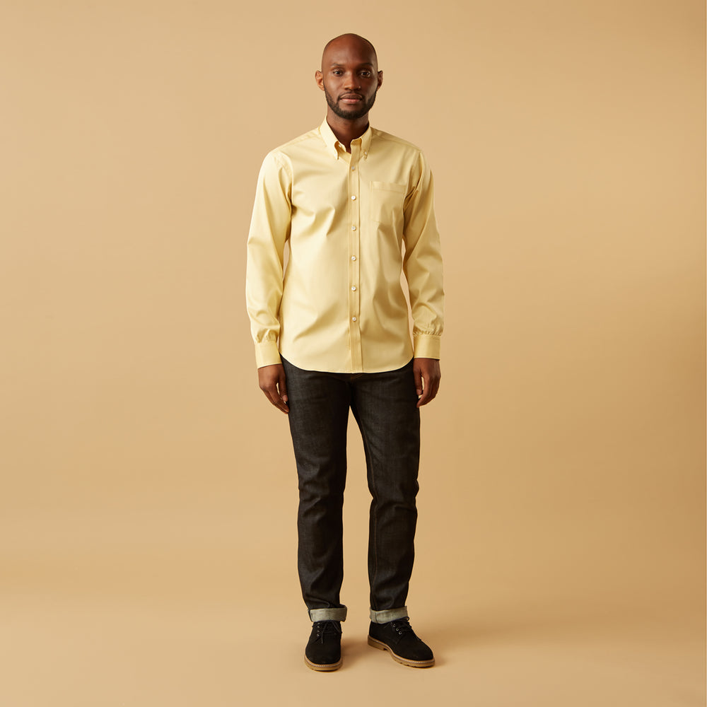 A man stands facing forward against a beige background, wearing the Turnbull & Asser Yellow Piece Dyed Weekend Fit Suffolk Shirt with a Dorset collar, paired with dark jeans and black shoes.