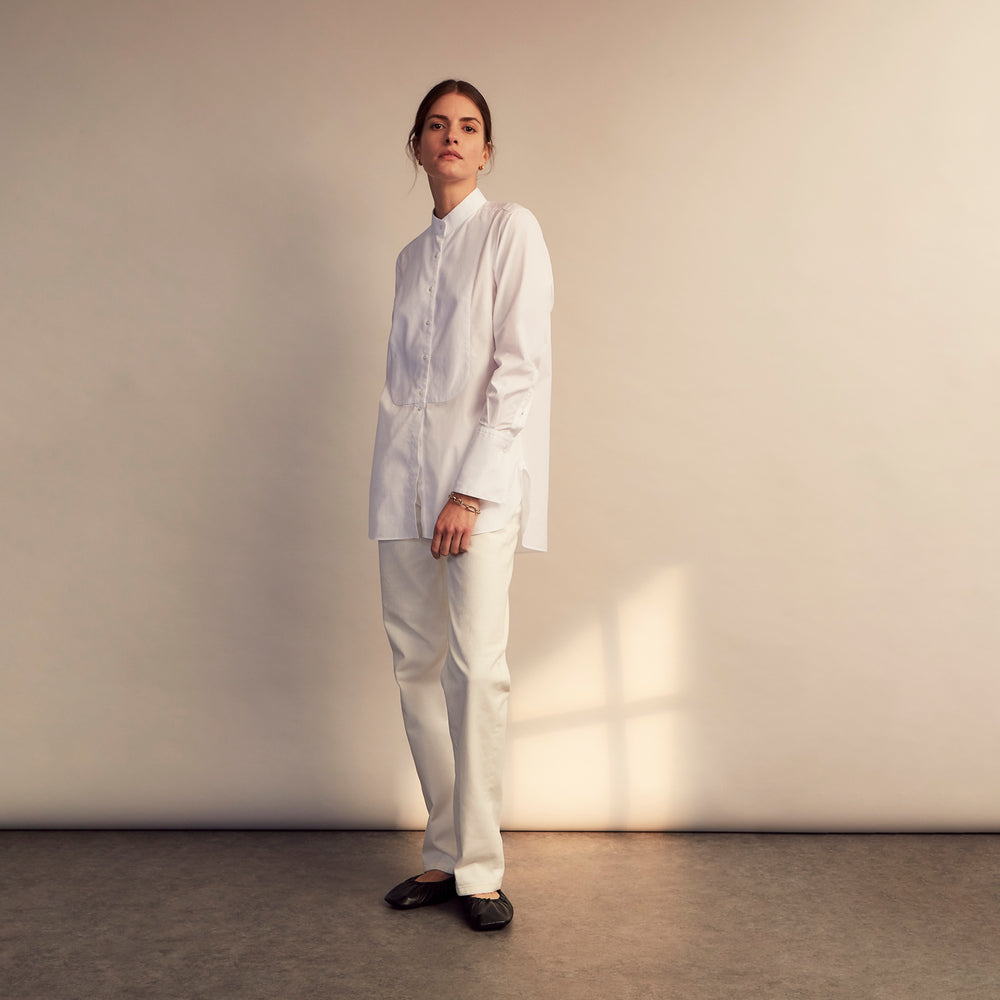 A person with curly hair wears the Turnbull and The Deck White Cotton Minimalist Shirt by Turnbull & Asser, paired with pants. Posed against a plain background, their minimalist silhouette is highlighted in this black and white image.