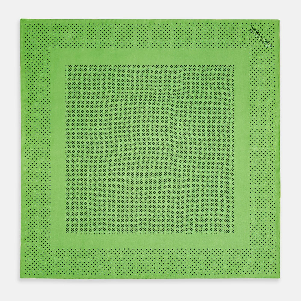 The Fern Green Dot Silk Pocket Square by Turnbull & Asser is crafted from pure silk, featuring a subtle black polka dot border with a denser dot pattern at the center and the brand name printed in the upper right corner.