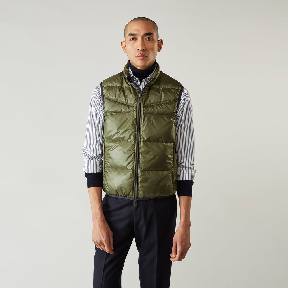 A man stands against a plain background wearing the Turnbull & Asser Green Peyton Sealup Quilted Gilet over a striped shirt, dark trousers, and white sneakers, with his hands in his pockets and looking slightly to the side.