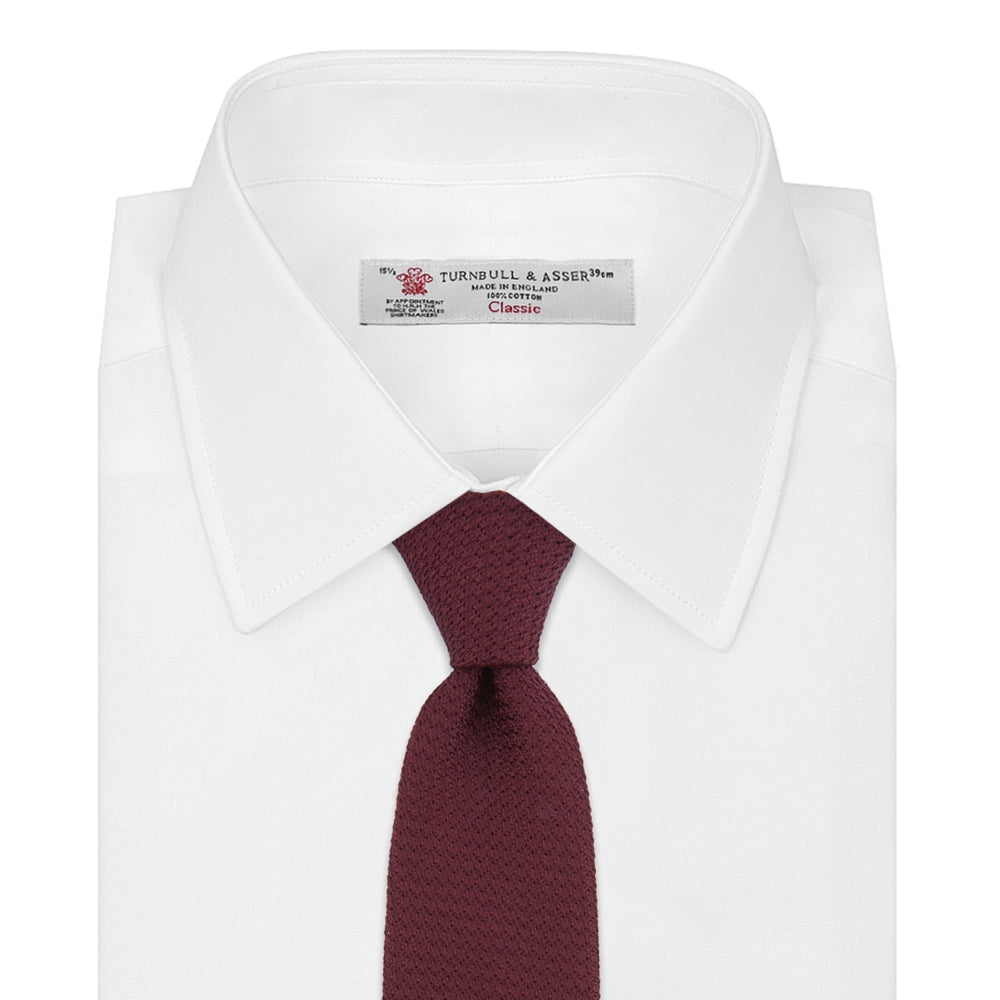 The Turnbull & Asser Plain Woven Grenadine Silk Tie, shown rolled at one end on a white background, features a rich burgundy hue and textured pattern—an essential accessory for sophisticated looks.