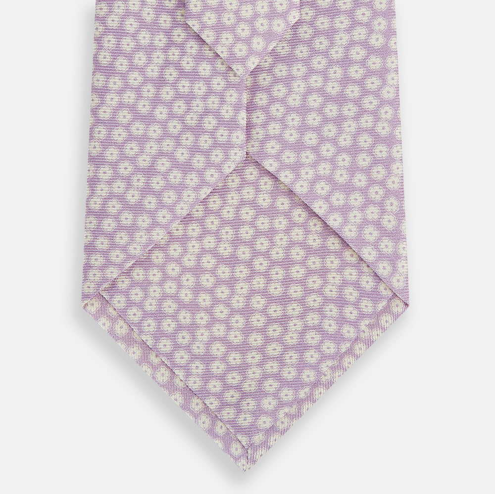 The Turnbull & Asser Lilac Flower-Patterned Printed Silk Tie, featuring a light lilac hue and small white circular designs, is shown rolled up against a plain white background.