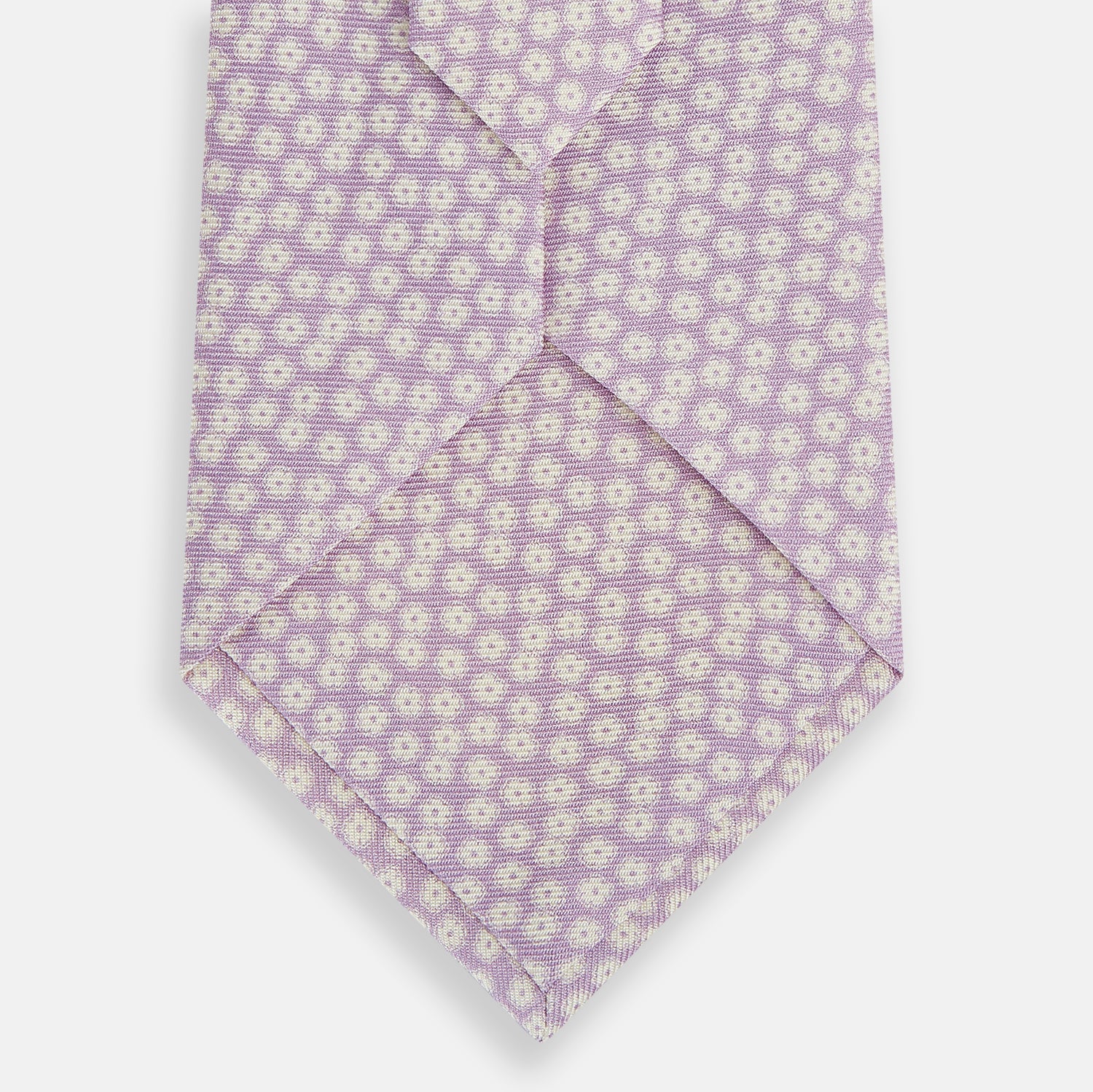 View 3 of product The Turnbull & Asser Lilac Flower-Patterned Printed Silk Tie, featuring a light lilac hue and small white circular designs, is shown rolled up against a plain white background.