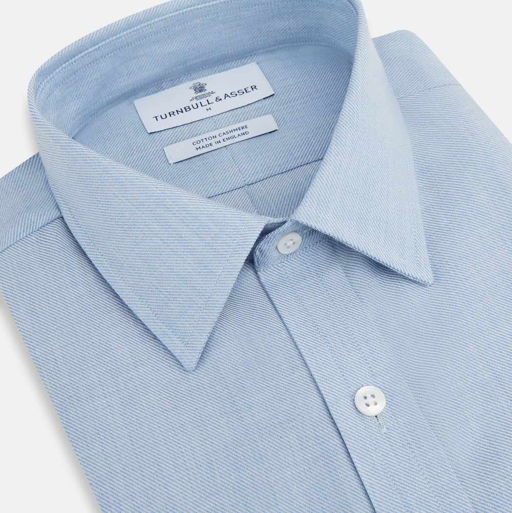 The Turnbull & Asser Pale Blue Cotton Cashmere Derby Collar Richmond Shirt features a soft cotton-cashmere blend, derby collar, white buttons, left chest pocket, and buttoned cuffs, shown laid flat on a white background.