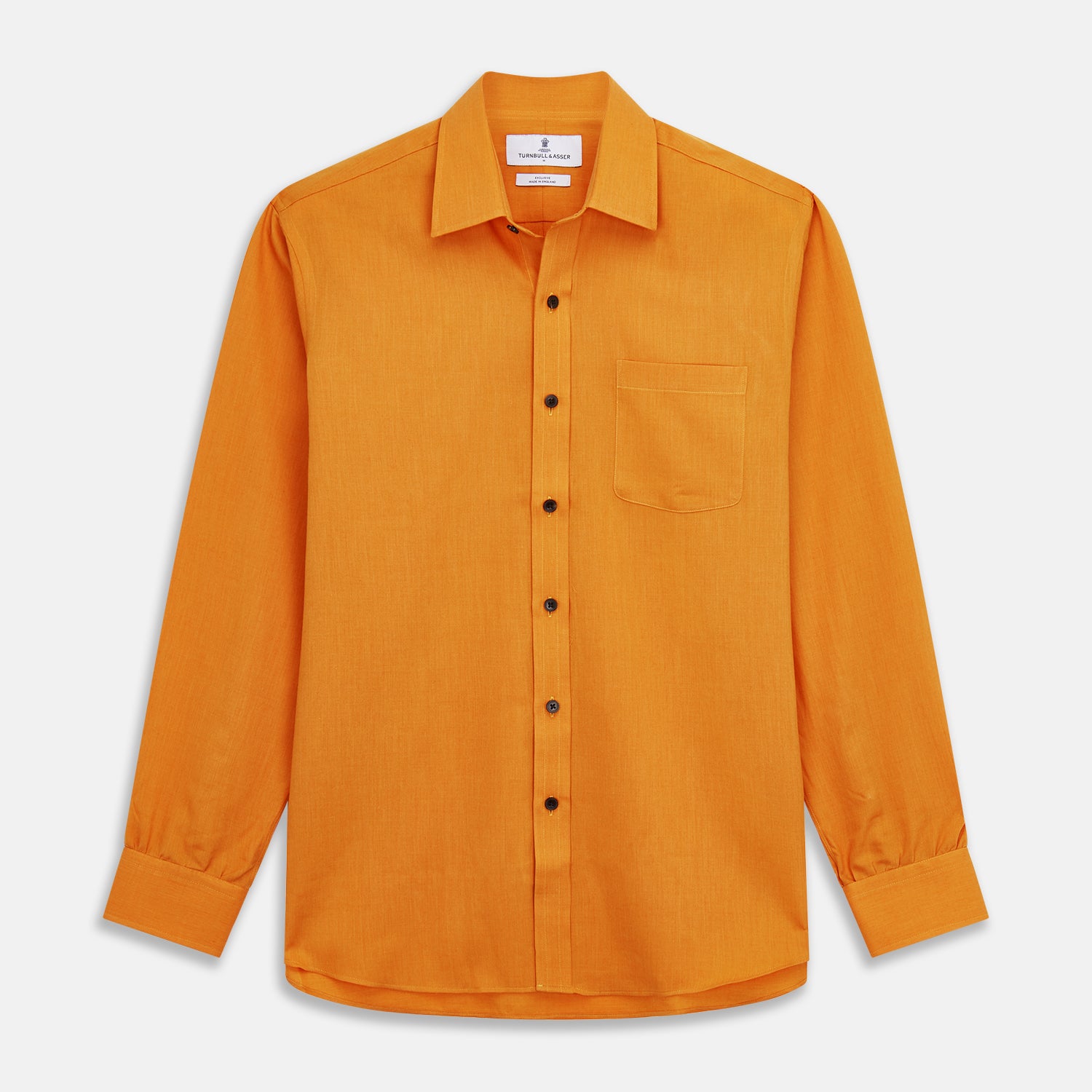 View 2 of product A man stands against a tan background, wearing a Turnbull & Asser Orange Weekend Fit Cotton & Wool Shirt with Derby Collar and 1 Button Cuffs, tucked into dark blue dress pants with a black belt and shoes. He has one hand in his pocket.