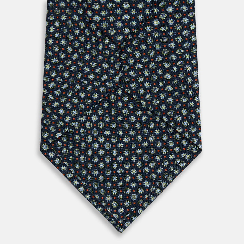 The Turnbull & Asser Navy Geometric Floral Silk Tie features a dark blue background with a repeating small geometric pattern in light blue, yellow, and red, shown rolled up on a plain white surface.