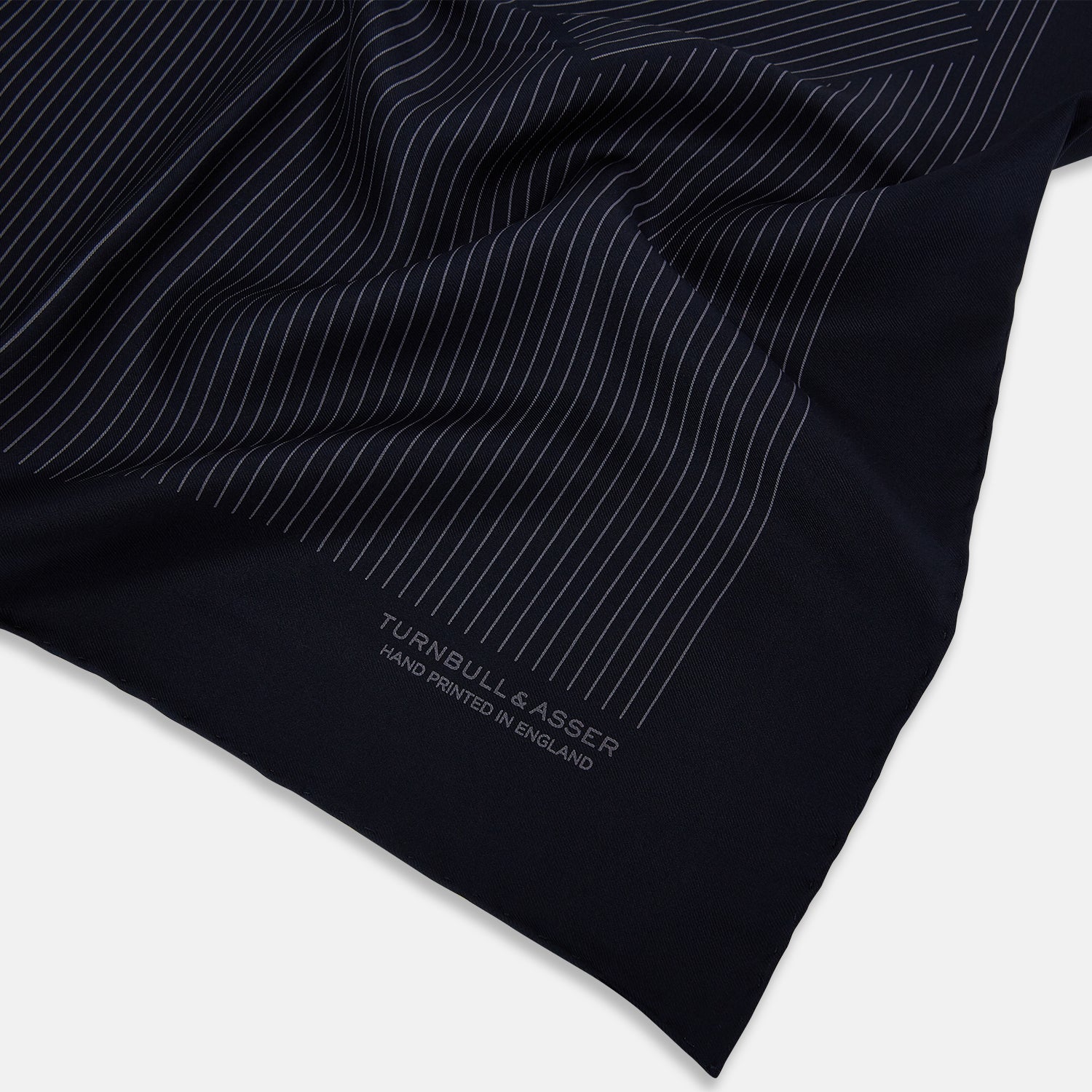 View 2 of product The Navy and Charcoal Stripe Silk Pocket Square by Turnbull & Asser features fine hand-rolled edges, thin white stripes on navy, and TURNBULL & ASSER HAND PRINTED IN ENGLAND in white on the corner.