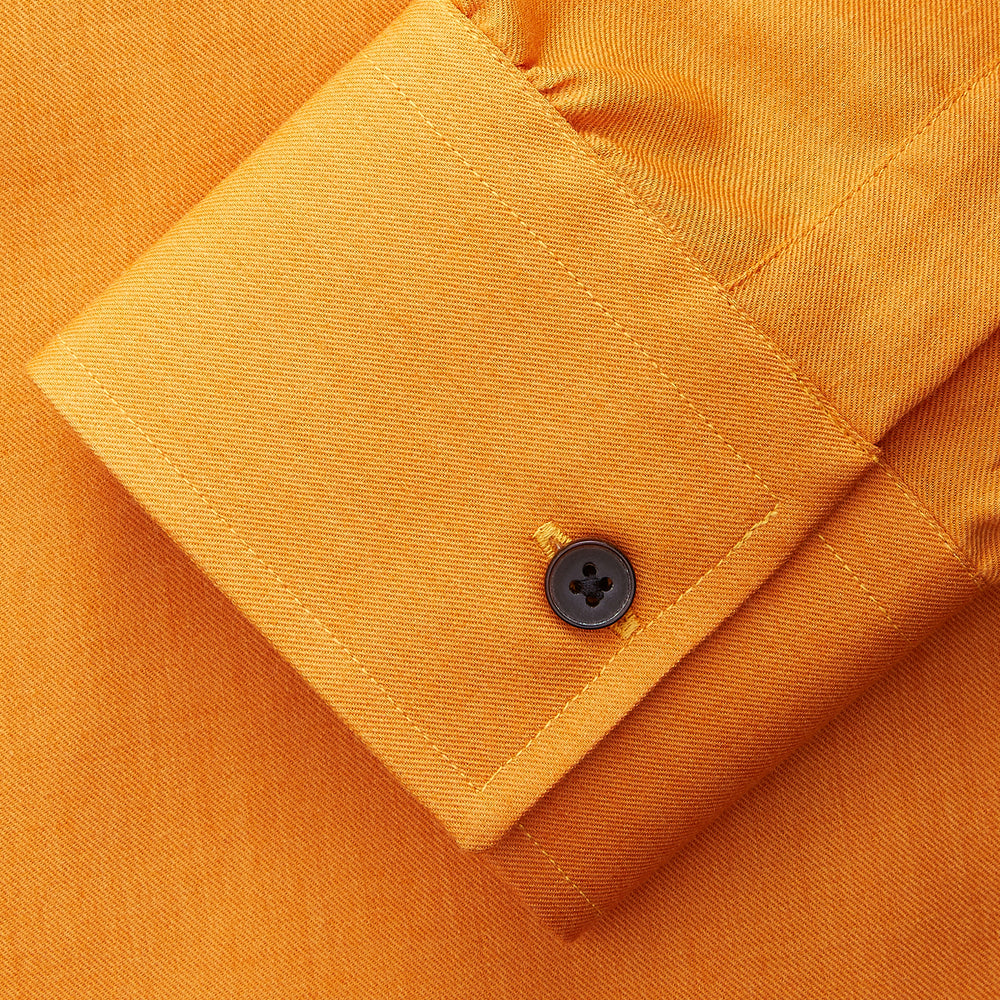 A man stands against a tan background, wearing a Turnbull & Asser Orange Weekend Fit Cotton & Wool Shirt with Derby Collar and 1 Button Cuffs, tucked into dark blue dress pants with a black belt and shoes. He has one hand in his pocket.
