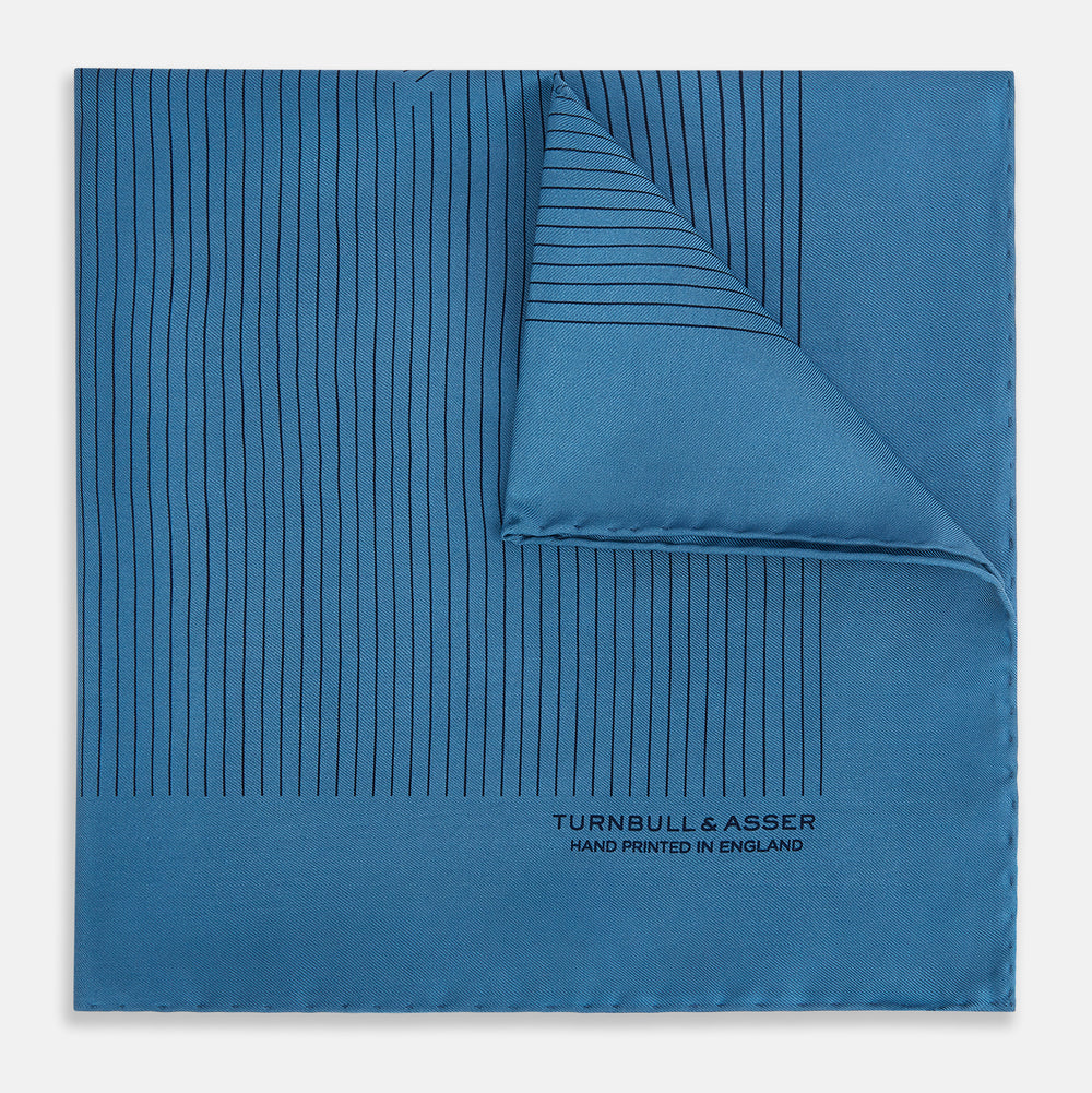 The Blue Stripe Silk Pocket Square by Turnbull & Asser features thin vertical lines, British heritage details, and "TURNBULL & ASSER HAND PRINTED IN ENGLAND" printed on the fabric, with one corner folded to reveal the reverse side.