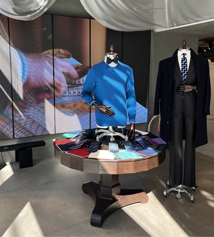 A stylish store display with two mannequins, one in a blue sweater and bow tie, the other in a dark suit and tie. A round table holds colorful fabrics and shoes. A large screen and coat are in the background.