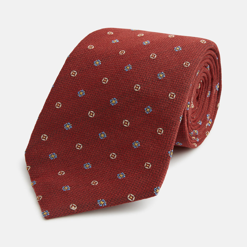 A Turnbull & Asser Red Motif Silk Tie featuring small blue and yellow floral designs with brown geometric shapes is displayed rolled up on a plain white background.