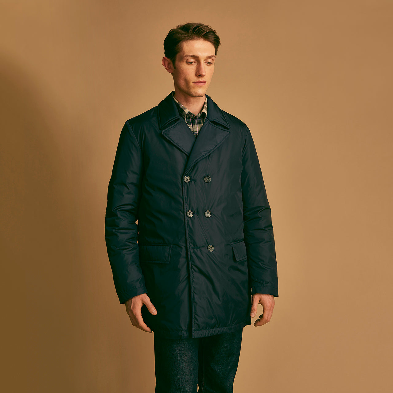 View 5 of product A man stands against a beige background, wearing the Turnbull & Asser Navy Burrows Double Breasted Coat over a plaid shirt and mauve cardigan, paired with dark rolled-up jeans and black shoes, looking directly at the camera with a neutral expression.