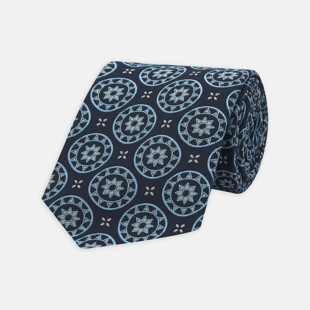 The Jacquard Silk Geometric Tie by Turnbull & Asser features a navy blue base with light blue and white star and circle motifs, shown rolled on a white background.