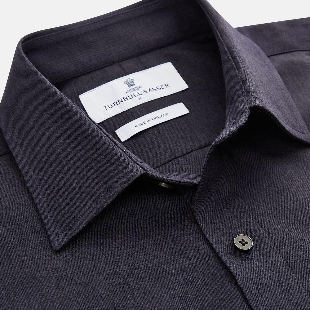 A man stands against a beige background wearing the Turnbull & Asser Navy Weekend Fit Cotton & Wool Shirt with a Derby collar and 1 button cuffs, paired with brown pants and black shoes, looking to the side with a neutral expression.