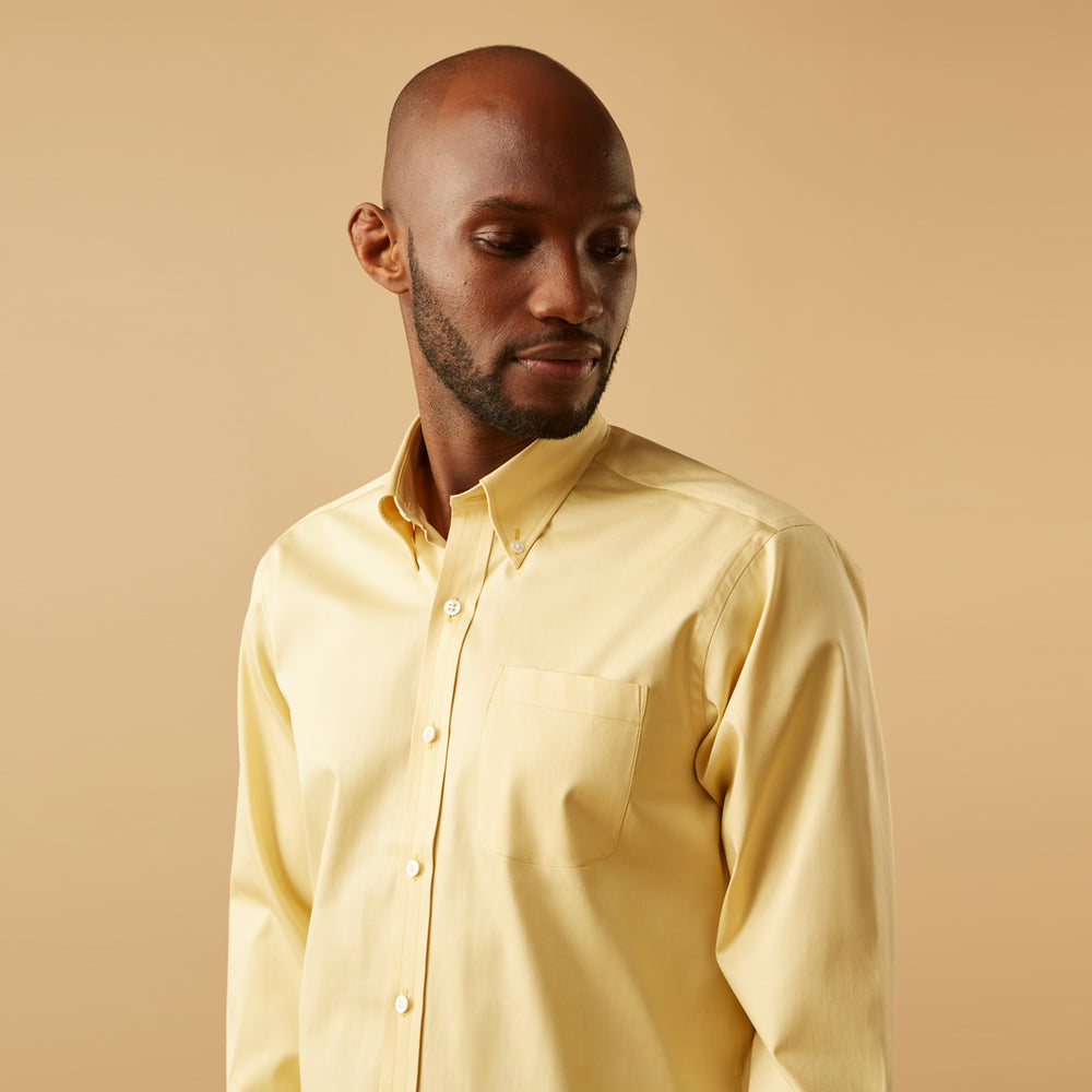 A man stands facing forward against a beige background, wearing the Turnbull & Asser Yellow Piece Dyed Weekend Fit Suffolk Shirt with a Dorset collar, paired with dark jeans and black shoes.