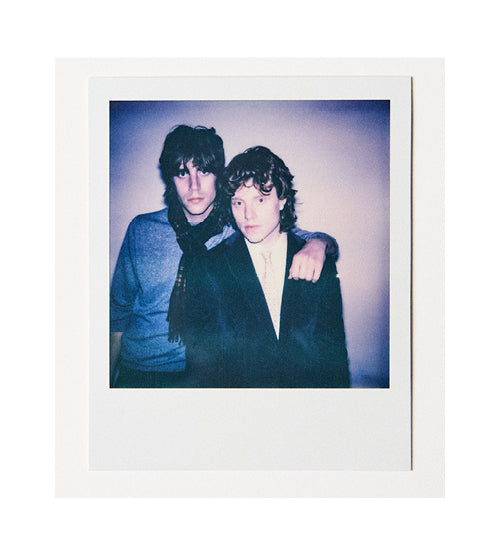 A Polaroid photo of two young people standing close together against a plain background. One has an arm around the other, and both have serious expressions. Their clothing is casual with one wearing a scarf.