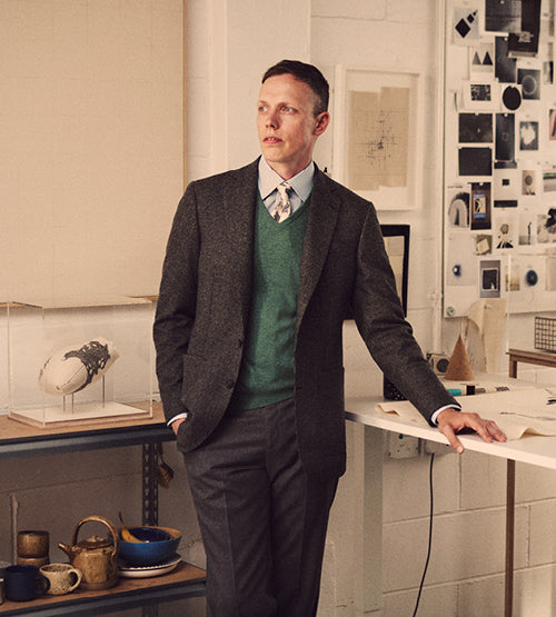 A man in a blazer, green sweater, and tie stands in an art studio, leaning on a table. Artworks, ceramics, and sketches are displayed on the wall and shelves around him.