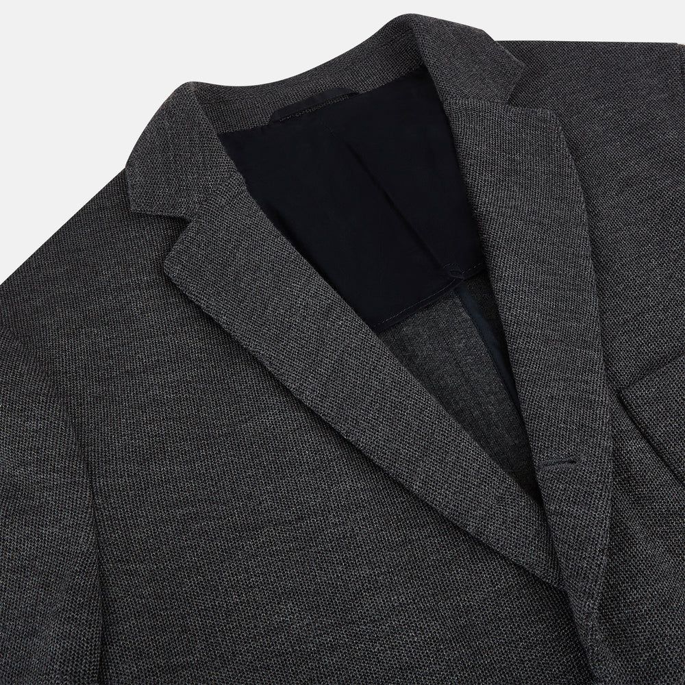 The Turnbull & Asser Graphite Knitted Oxford Blazer for men features a dark gray hue, two buttons, notched lapels, and three front pockets—a modern British-inspired piece styled elegantly against a light background.