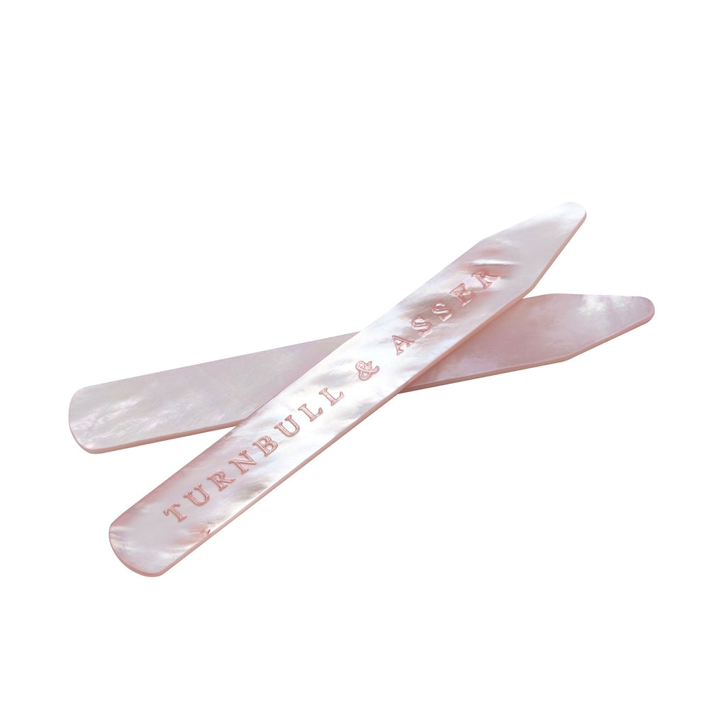 Two Pink Mother-of-Pearl Collar Stays from Turnbull & Asser rest on a white surface; one stay features the brand engraved in pink lettering, making them refined accessories for shirts.