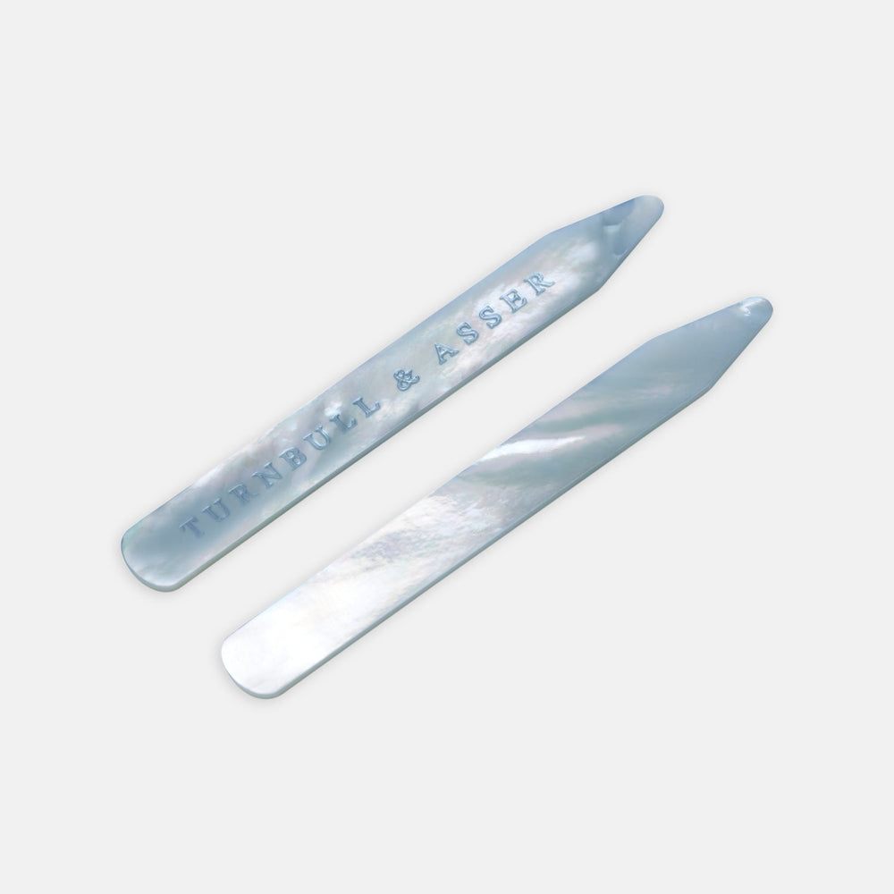 Two blue mother-of-pearl collar stays by Turnbull & Asser, ideal for keeping your shirt collar crisp; one is engraved with the brand name and both are displayed on a light gray background.