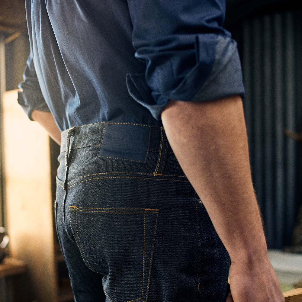 A pair of Turnbull & Asser Indigo Japanese Selvedge Denim Jeans in dark blue with rolled-up cuffs, yellow stitching, five pockets, and a button fly, laid flat on a white background.