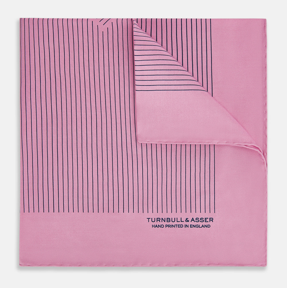 The Turnbull & Asser Pink Stripe Silk Pocket Square features thin vertical navy stripes and "TURNBULL & ASSER HAND PRINTED IN ENGLAND" in the bottom right corner, with one corner stylishly folded over.