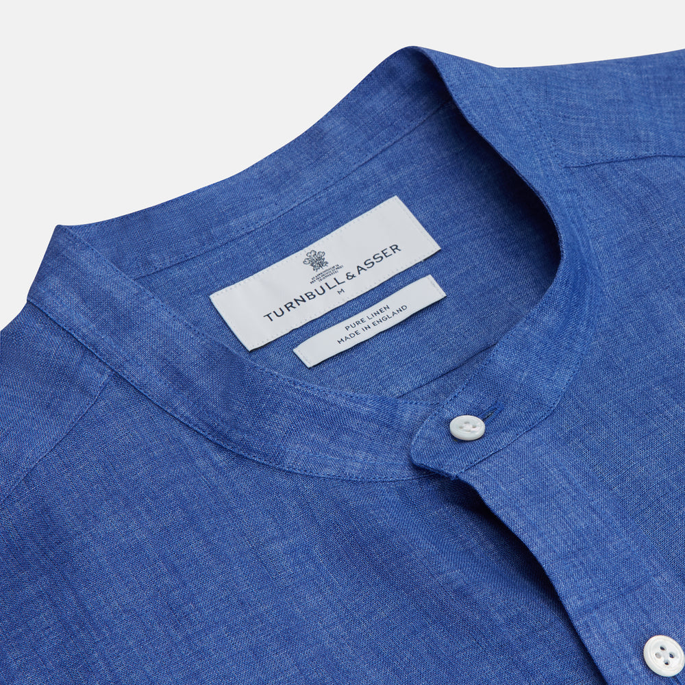A man faces forward with a neutral expression, hands in his pockets, wearing the Turnbull & Asser Plain Indigo Linen Weekend Fit Shirt with a stand collar, styled with dark pants and black shoes against a plain light background.