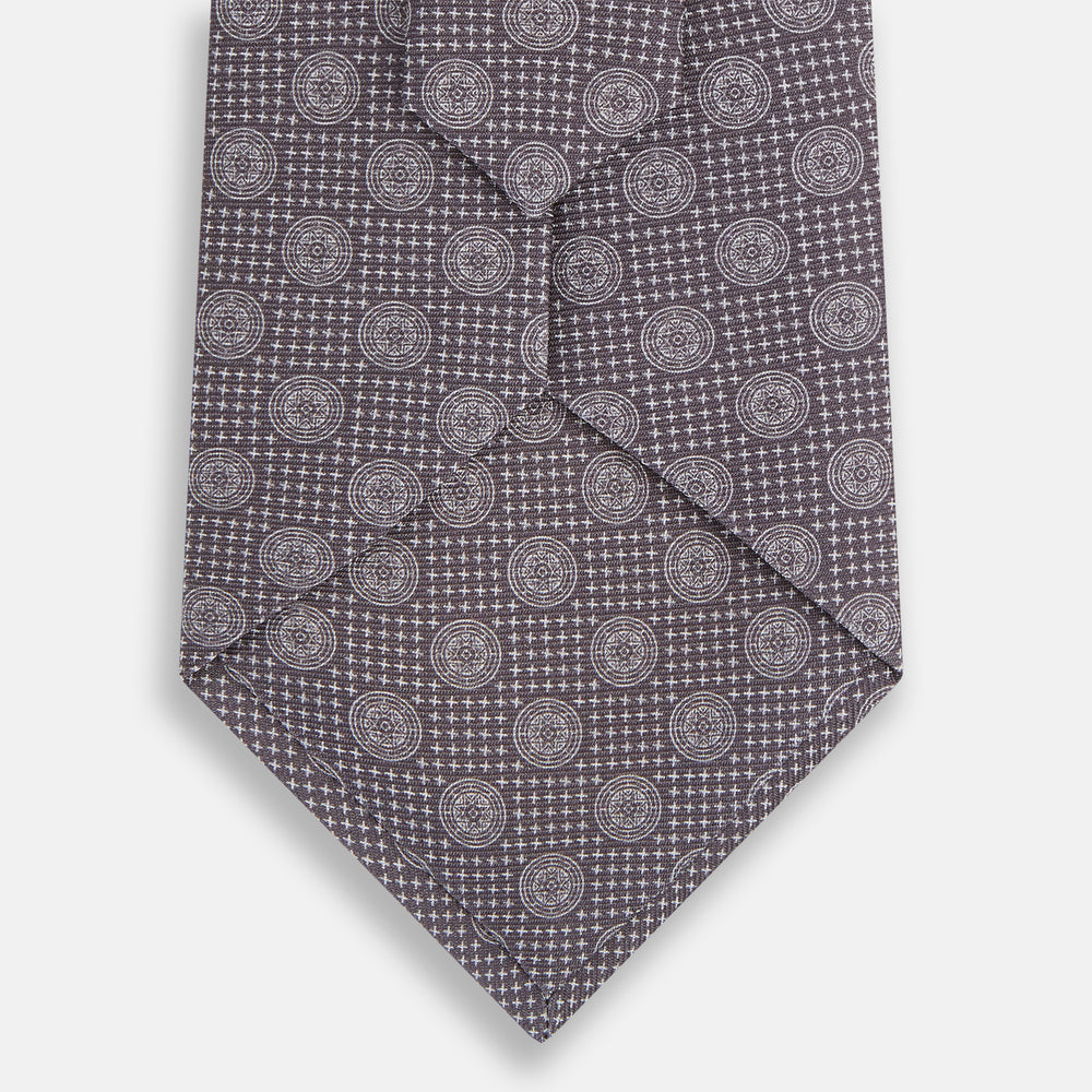 The Turnbull & Asser Ash Medallion Silk Tie is shown rolled up on a white background. This purple tie features a repeating geometric motif with small white circles and grid-like designs.