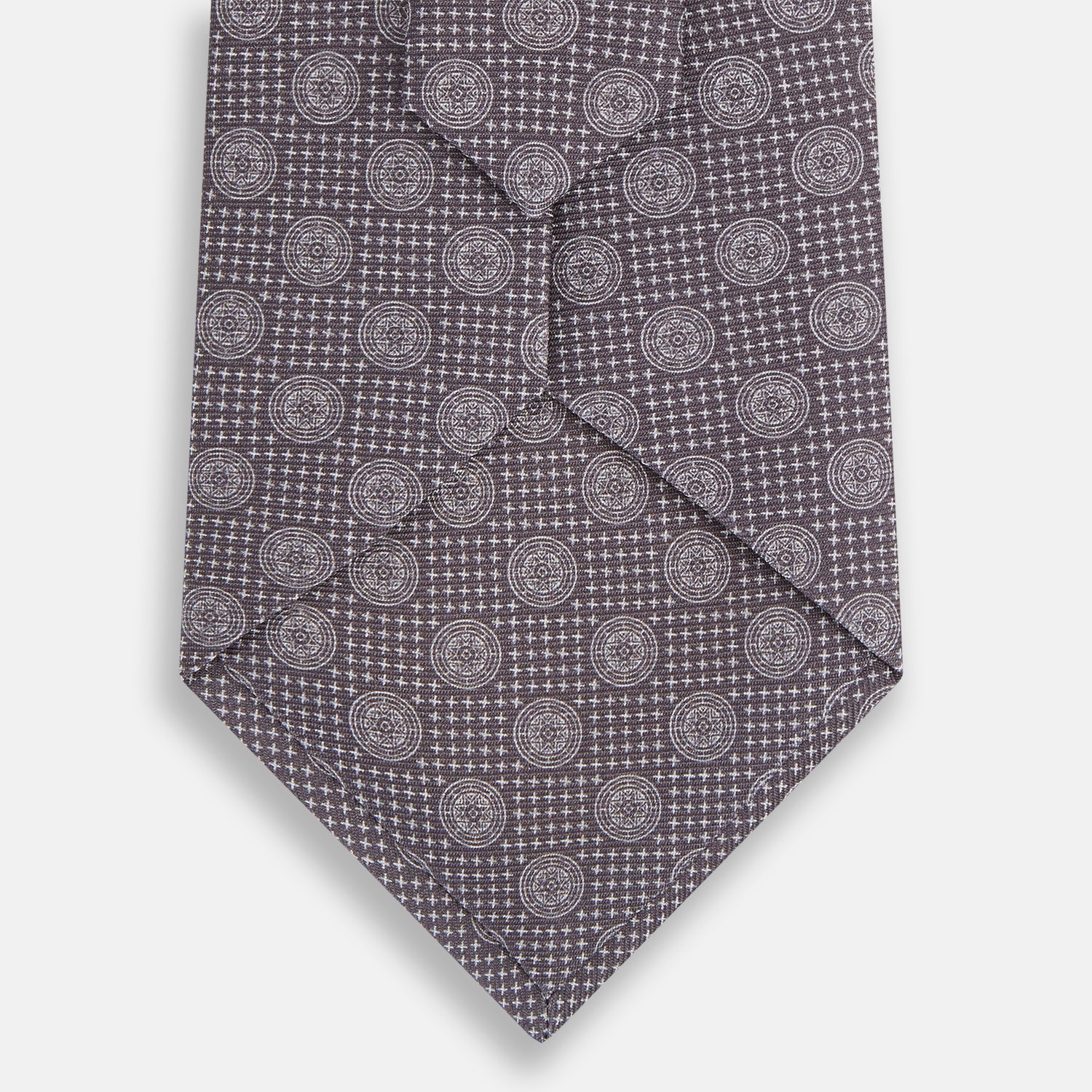 View 3 of product The Turnbull & Asser Ash Medallion Silk Tie is shown rolled up on a white background. This purple tie features a repeating geometric motif with small white circles and grid-like designs.