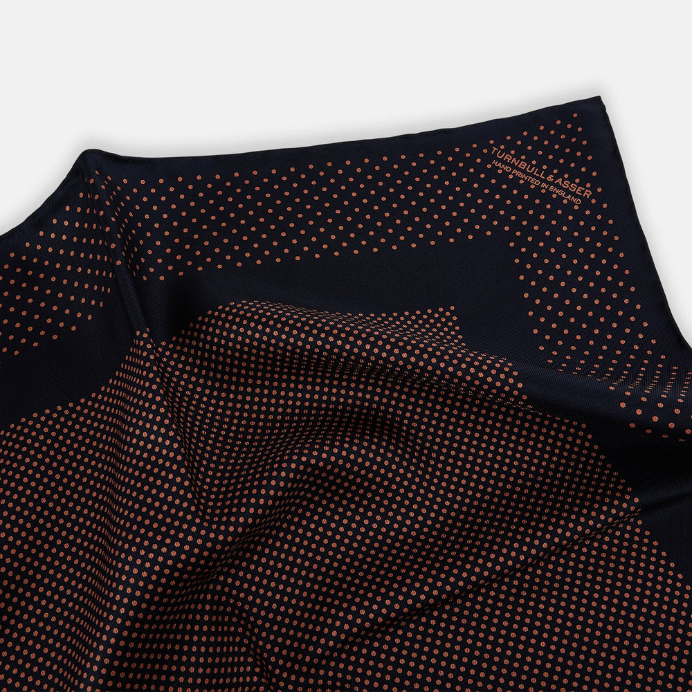 The Navy and Blush Dot Silk Pocket Square by Turnbull & Asser features a navy base with small orange polka dots and a border, shown neatly folded with one corner flipped over.